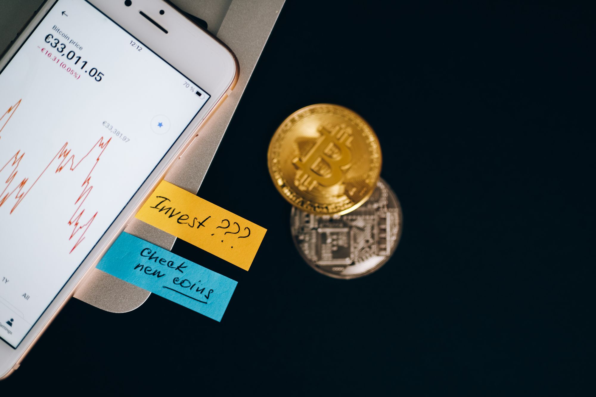 A phone screen with bitcoin price on it next to two paper strips, a bitcoin and another coin.