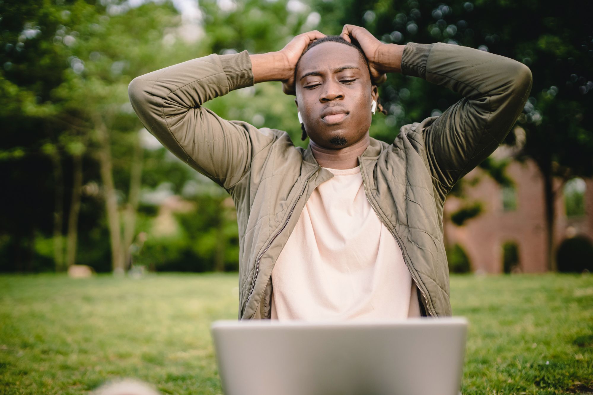 A man sitting on grass with a laptop on his lap and his hands on his head